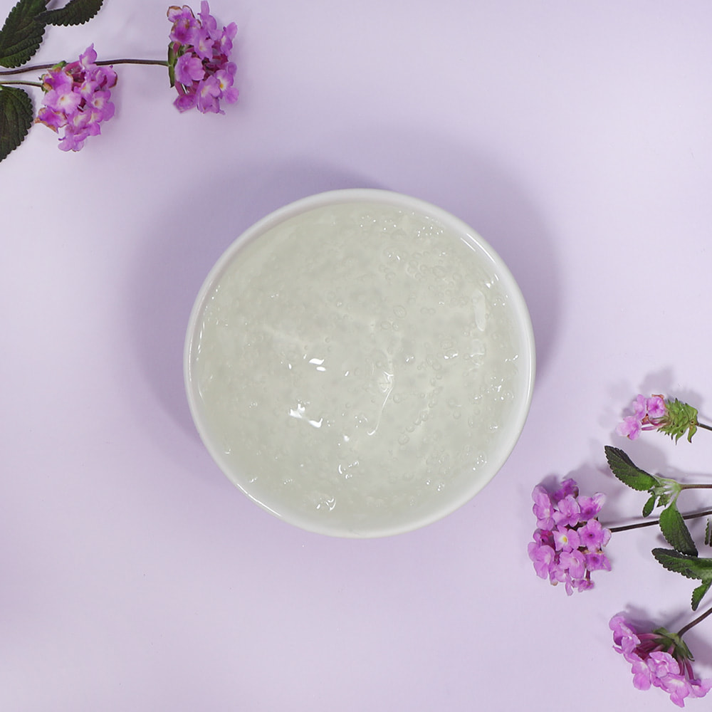 A top-down view of a white bowl filled with a clear, bubbly suspension shower gel base, gracefully framed by delicate purple flowers and green leaves on a pastel purple background.