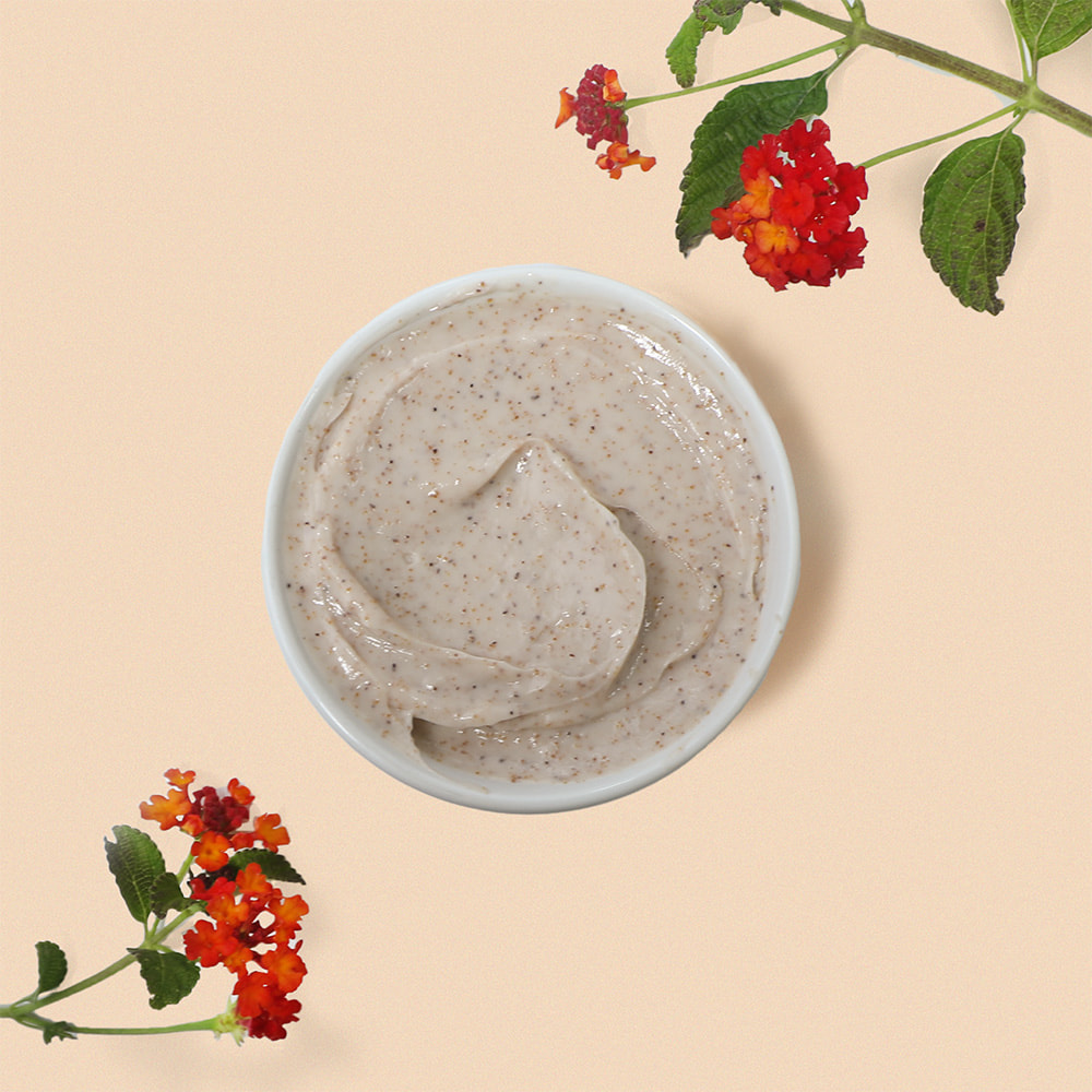 A top-down view of a white bowl filled with a creamy body scrub speckled with finely milled walnut shells, elegantly framed by vibrant red flowers and green leaves on a pastel peach background.
