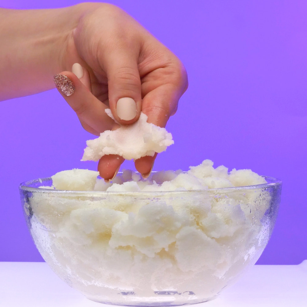 A close-up of a person's fingers pinching and lifting a thick, moldable clump of bright white sugar scrub from a clear glass bowl against a vibrant purple background.
