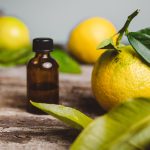 A small amber glass dropper bottle resting on a rustic, textured wooden surface, surrounded by fresh, whole yellow-green bergamot citrus fruits and vibrant green leaves.