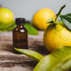 A small amber glass dropper bottle resting on a rustic, textured wooden surface, surrounded by fresh, whole yellow-green bergamot citrus fruits and vibrant green leaves.