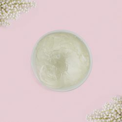 A top-down view of a white bowl filled with chunky, transparent jelly soap base, framed by delicate white flowers in the corners against a pastel pink background.