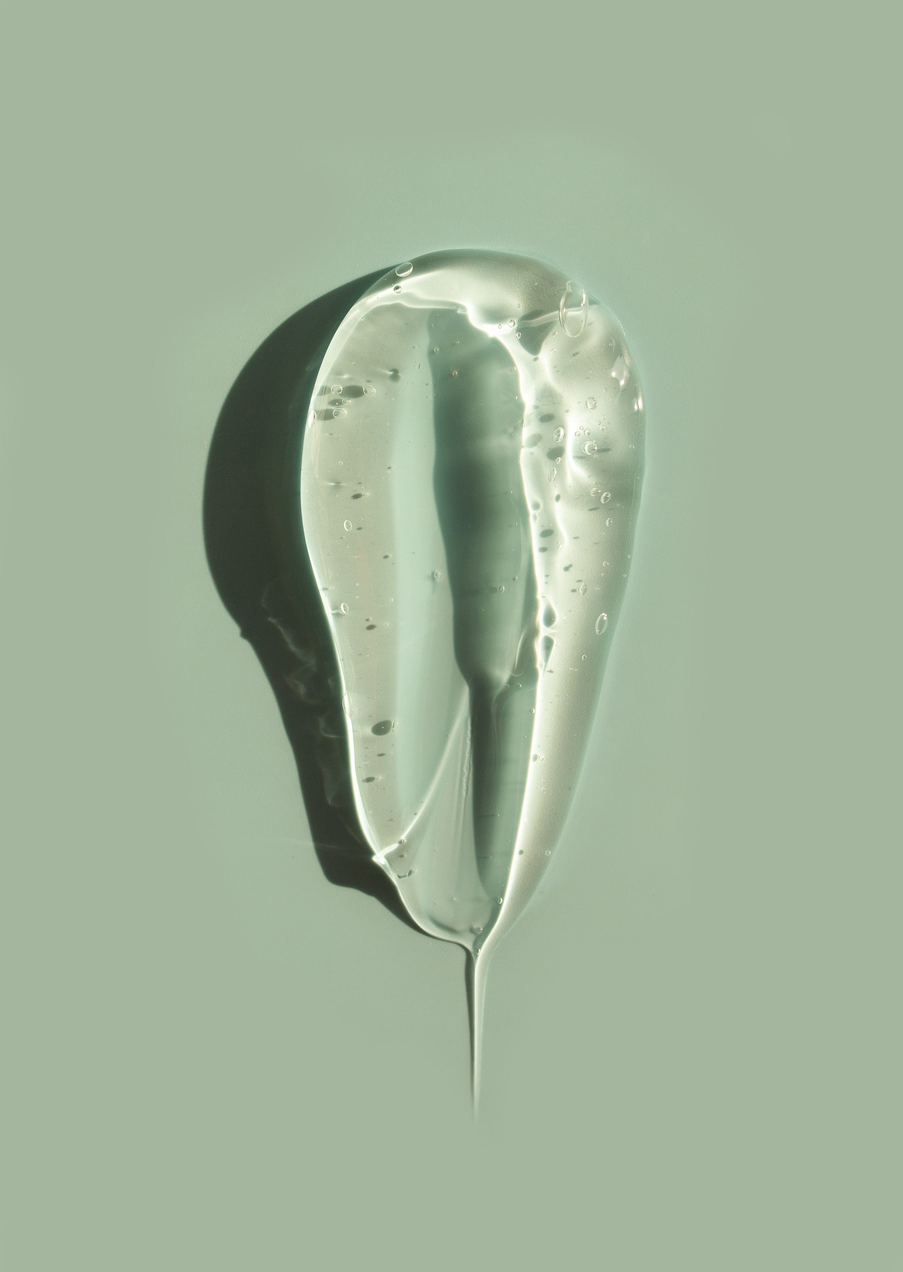A thick, transparent swatch of clear aloe vera gel showing small suspended air bubbles and a glossy finish against a soft green background.