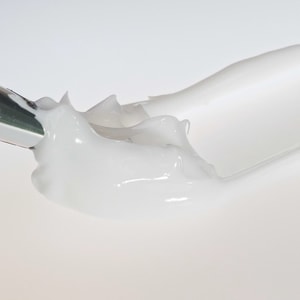 A close-up macro shot of a thick, glossy white cosmetic lotion base being smoothly smeared across a white surface by a metal mixing spatula.