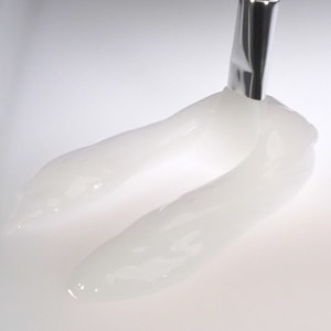 A close-up shot of a clear, semi-viscous aftershave gel being smoothly smeared across a white surface by a metal cosmetic mixing spatula.