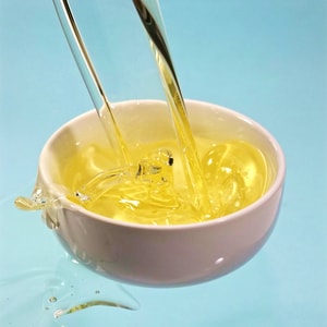 Clear, pale yellow massage oil being poured into a white ceramic bowl from two different streams, creating dynamic ripples and splashes against a bright blue background.