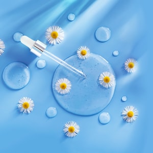 A clear, liquid puddle of hair spray base with a glass dropper resting inside, surrounded by fresh white chamomile flowers and small water droplets on a bright blue background.