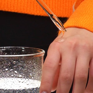 A close-up of a clear glass dropper dripping a thick, transparent drop of Argan hair serum onto the back of a hand, next to a condensation-covered glass of water.