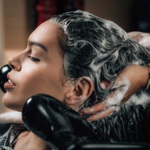 A close-up profile of a woman relaxing at a salon basin with her eyes closed as a stylist massages a thick, white lather of shampoo into her wet, dark hair.