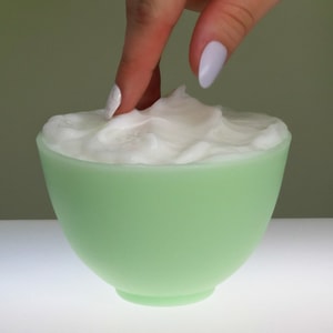 A close-up shot of two fingers gently scooping thick, bright white lotion from a small, soft green bowl.
