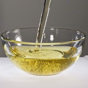 A stream of clear, golden yellow oil being poured into a transparent glass bowl, creating a pool of liquid with small air bubbles against a grey and white background.