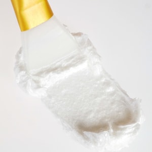 A close-up macro shot of a thick, pearlescent white facial cleanser being smoothly smeared across a white surface by a white silicone spatula with a gold handle.