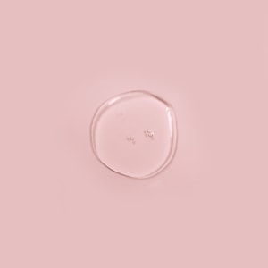A top-down view of a clear, perfectly transparent puddle of liquid soap with small bubbles resting gracefully on a solid soft pink background.