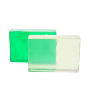 Two smooth blocks of transparent melt and pour soap, one tinted bright green and the other uncolored, standing together against a crisp white background.