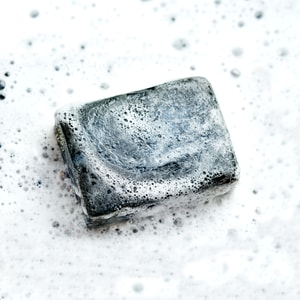 A wet bar of dark grey charcoal soap covered in a thick, rich white foam lather against a bubbly white background.