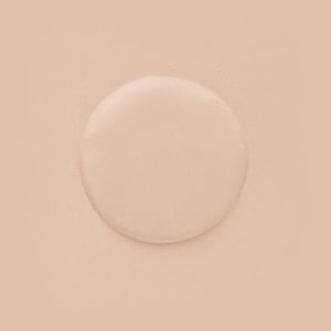 A top-down view of a single, perfectly round puddle of clear liquid resting on a lightly textured, beige surface.