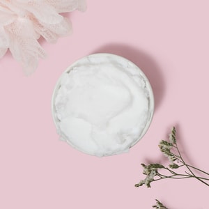 A top-down view of a white bowl filled with a thick, fluffy white foaming bath whip base, gracefully framed by a pink shower loofah and delicate flowers on a pastel pink background.