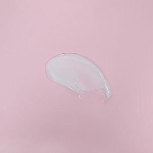 A top-down close-up of a smooth, thick swatch of glossy white hair conditioner against a solid soft pink background.