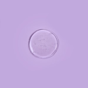 A top-down close-up of a clear, perfectly round puddle of liquid shampoo with tiny suspended bubbles resting on a solid pastel purple surface.