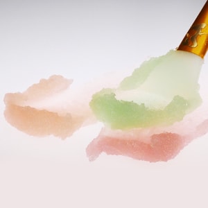 A close-up of a gold-handled spatula smearing three distinct swatches of sugar scrub in pastel peach, green, and pink across a crisp white surface.