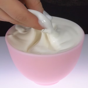 A close-up shot of a hand gently pinching and lifting a smooth, glossy white facial cream upward from a soft pink bowl, showing its elegant consistency.