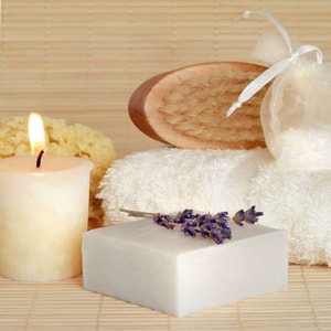 A tranquil spa setting featuring a square white bar of soap topped with dried lavender, placed next to a glowing lit candle, folded white towels, and a wooden bath brush on a bamboo mat.