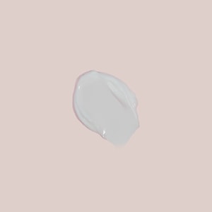 A top-down close-up of a smooth, glossy swatch of white facial cream resting on a solid soft pink surface.
