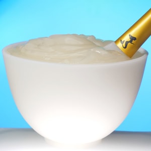 A close-up shot of a white bowl filled with thick, glossy, translucent gel cream, with a gold-handled spatula resting inside it against a vibrant blue background.