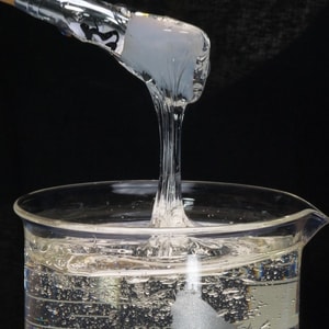 A close-up shot of a tool lifting a thick, stringy ribbon of highly viscous, clear liquid glycerin out of a glass measuring beaker against a stark black background.