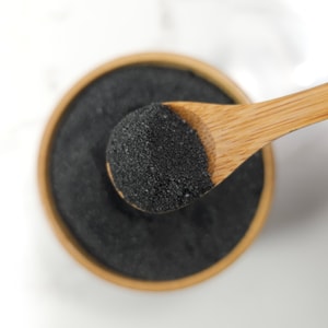 A top-down close-up of a wooden bamboo spoon lifting a scoop of fine, black Dead Sea salt crystals out of a matching bamboo bowl against a smooth white background.