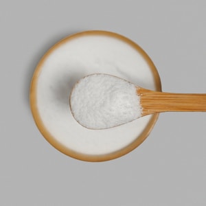 A top-down close-up of a wooden bamboo spoon lifting a scoop of fine, white foaming bath salts over a matching bamboo bowl against a light grey background.