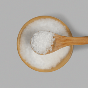 A top-down close-up of a wooden bamboo spoon lifting a scoop of medium, white foaming bath salts over a matching bamboo bowl against a light grey background.