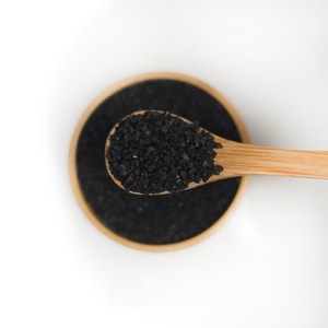 A top-down close-up of a wooden bamboo spoon lifting a scoop of small, black sea salt crystals out of a matching bamboo bowl against a smooth white background.