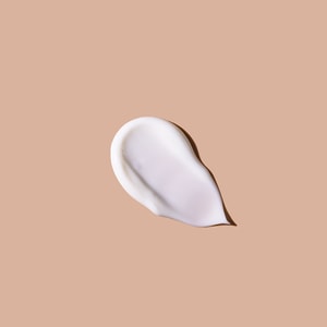A top-down close-up of a smooth, glossy swatch of thick white body custard resting on a solid soft tan surface.