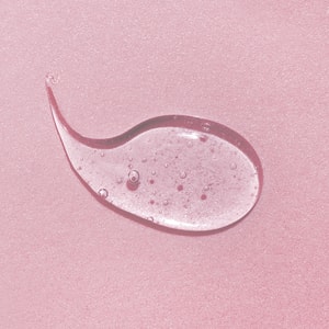 A top-down close-up of a perfectly clear, semi-viscous swatch of liquid soap base featuring tiny suspended bubbles, resting on a textured pastel pink surface.