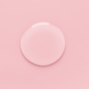 A top-down close-up of a perfectly clear, round puddle of liquid room spray base resting on a solid pastel pink surface.