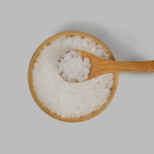 A top-down close-up of a wooden bamboo spoon lifting a scoop of coarse, white sea salt crystals out of a matching bamboo bowl against a light grey background.