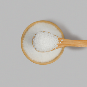 A top-down close-up of a wooden bamboo spoon lifting a scoop of medium-sized, white sea salt crystals out of a matching bamboo bowl against a light grey background.
