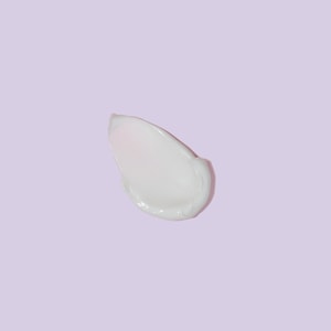 A top-down close-up of a smooth, glossy swatch of white shaving lotion resting on a solid pastel purple surface.