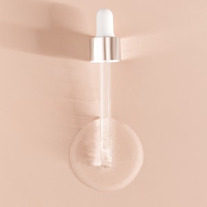 A top-down close-up of a white and silver glass dropper resting inside a perfectly round puddle of clear, lightweight dry oil on a solid peach surface.