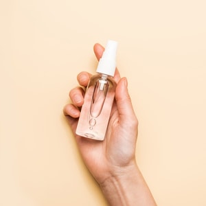 A close-up of a hand holding a small, clear plastic pump spray bottle filled with a transparent rinse-free hand wash against a solid peach background.