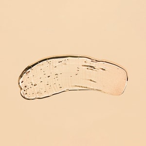 A top-down close-up of a thick, transparent swatch of hair styling gel featuring suspended bubbles, resting on a solid light peach surface.