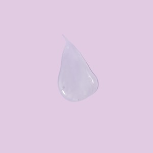 A top-down close-up of a perfectly clear, smooth droplet of thick liquid bubble bath resting on a solid pastel purple surface.