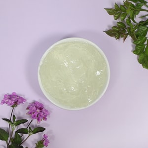 A top-down view of a white bowl filled with a thick, clear hair styling gel base, gracefully framed by purple flowers and green leaves on a pastel purple background.
