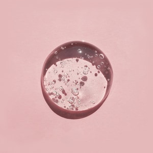 A top-down close-up of a perfectly clear, thick swatch of suspension shower gel packed with suspended air bubbles, casting a distinct shadow on a solid pink surface.