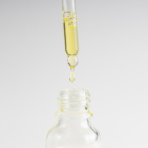 A close-up of a glass dropper dispensing a single drop of clear, pale yellow dry oil into the neck of a clear glass bottle against a clean white background.