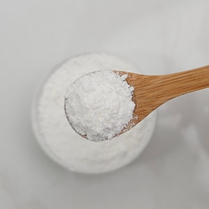 A top-down close-up of a wooden bamboo spoon holding a scoop of dry, white fizzy bath powder against a blurred light grey background.