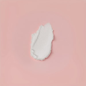 A top-down close-up of a thick swatch of white foaming cleansing scrub, showing its dense, slightly textured consistency against a solid soft pink background.