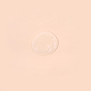 A top-down close-up of a perfectly round, clear puddle of liquid wash base featuring tiny suspended bubbles, resting on a solid pastel peach surface.