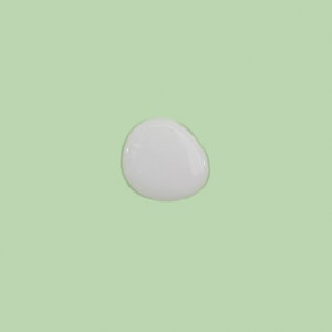 A top-down close-up of a perfectly round, creamy white puddle of liquid wash base resting on a solid pastel green surface.